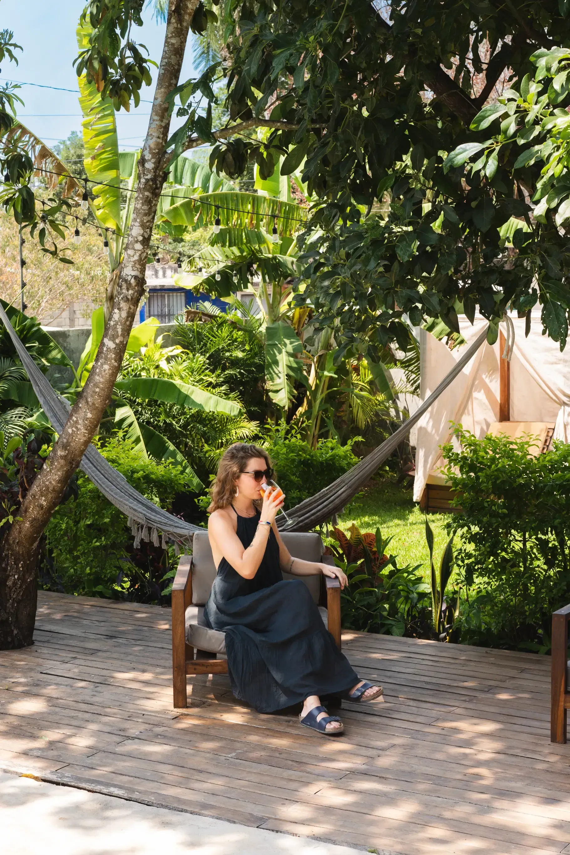 Una mujer con gafas de sol bebe una bebida en un salón al aire libre, rodeada de vegetación y una hamaca.