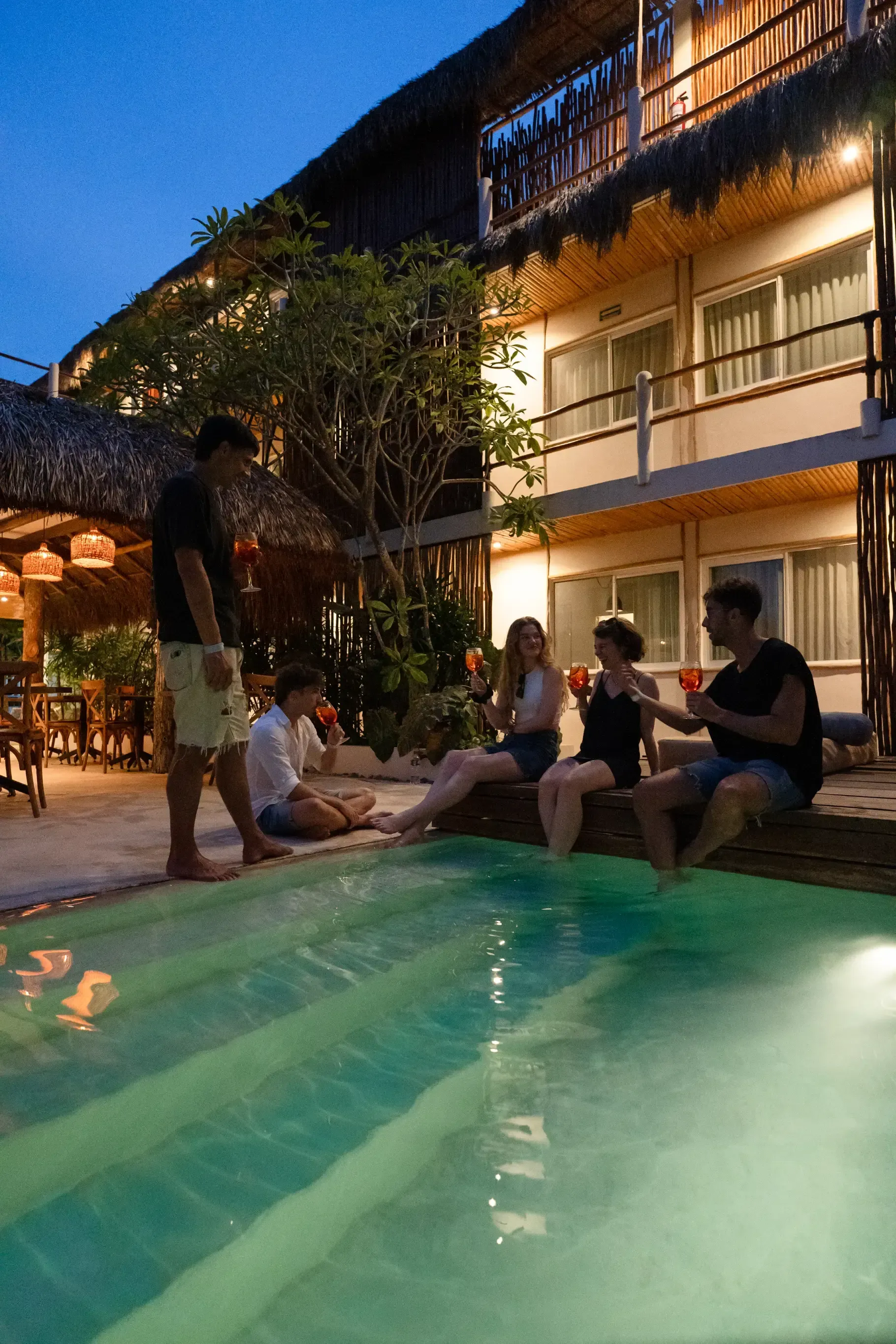 Amigos brindando con bebidas alrededor de una piscina por la noche cerca de un resort de estilo tropical.