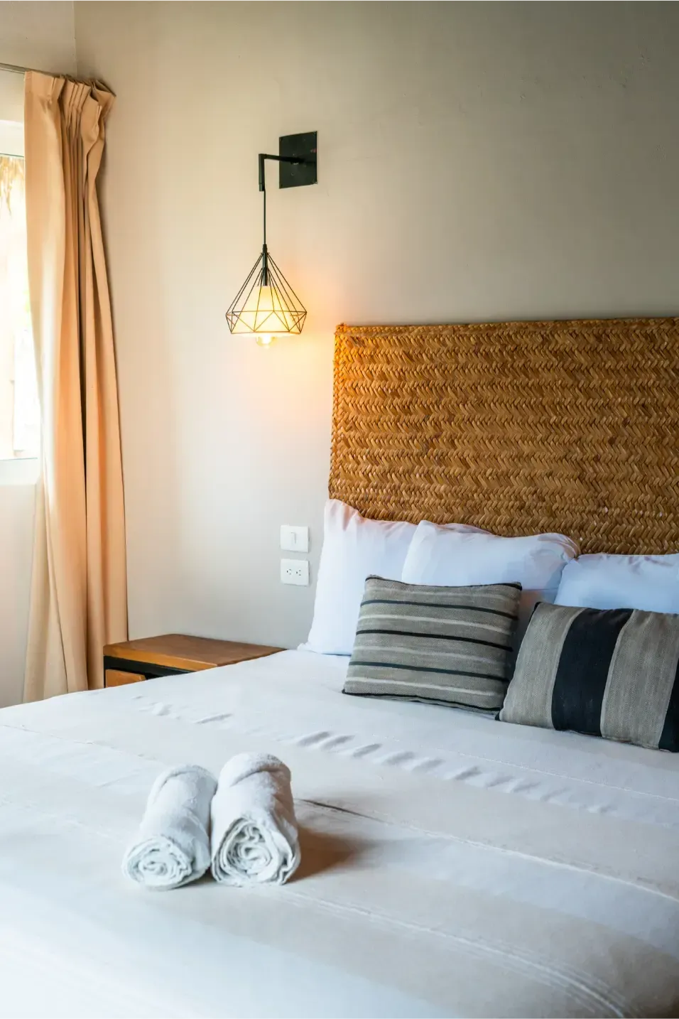 Dormitorio con cama, cabecero tejido, almohadas, toallas enrolladas y lámpara decorativa.