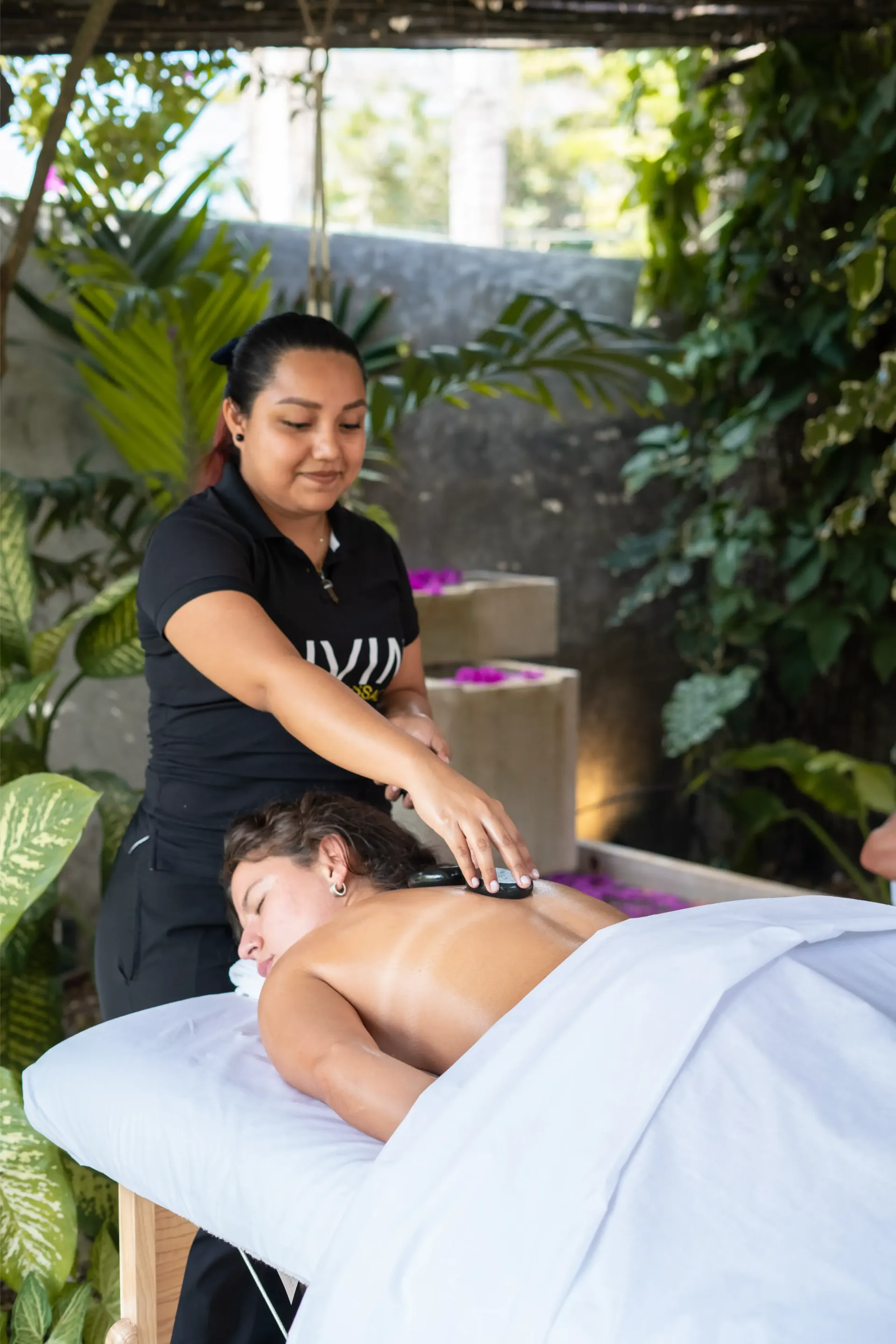 Mujer recibiendo masaje con piedras calientes al aire libre; terapeuta con top negro, follaje verde.