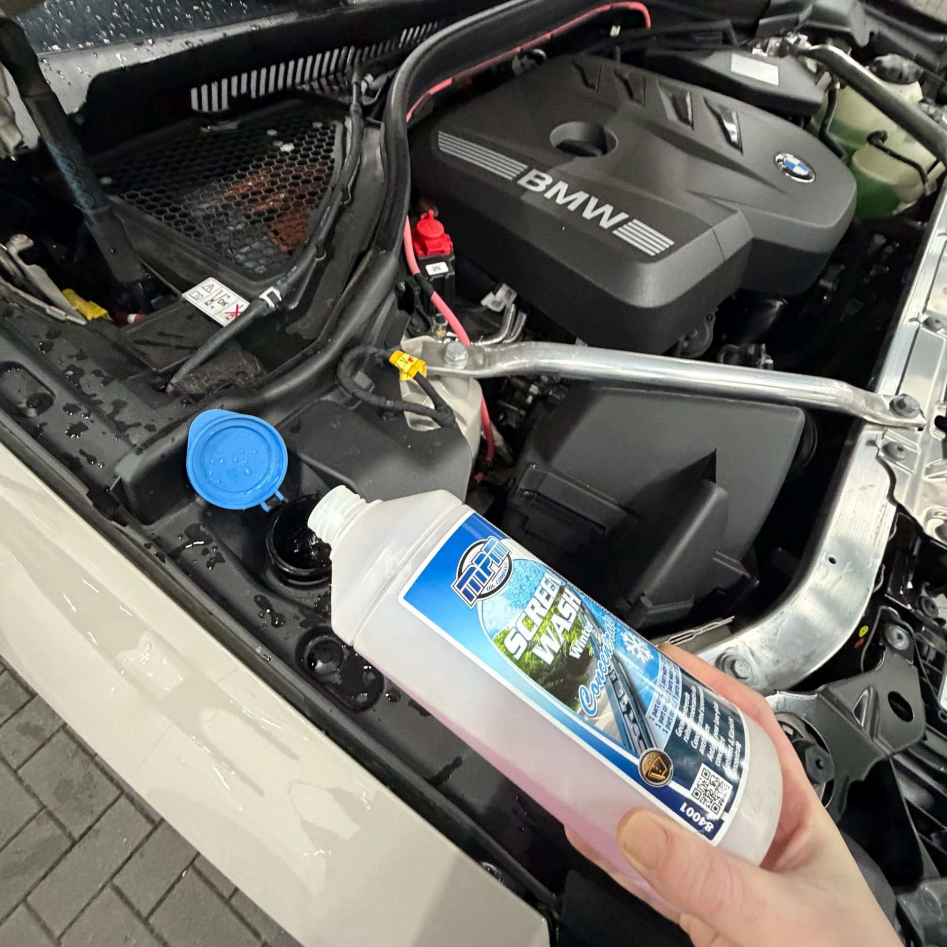 Hand die een fles ruitenwisservloeistof vasthoudt in de buurt van de motor van een BMW.