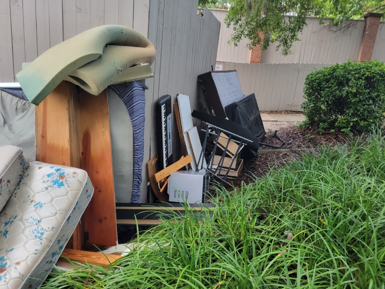 Pile of discarded furniture and debris next to a fence and overgrown bushes.