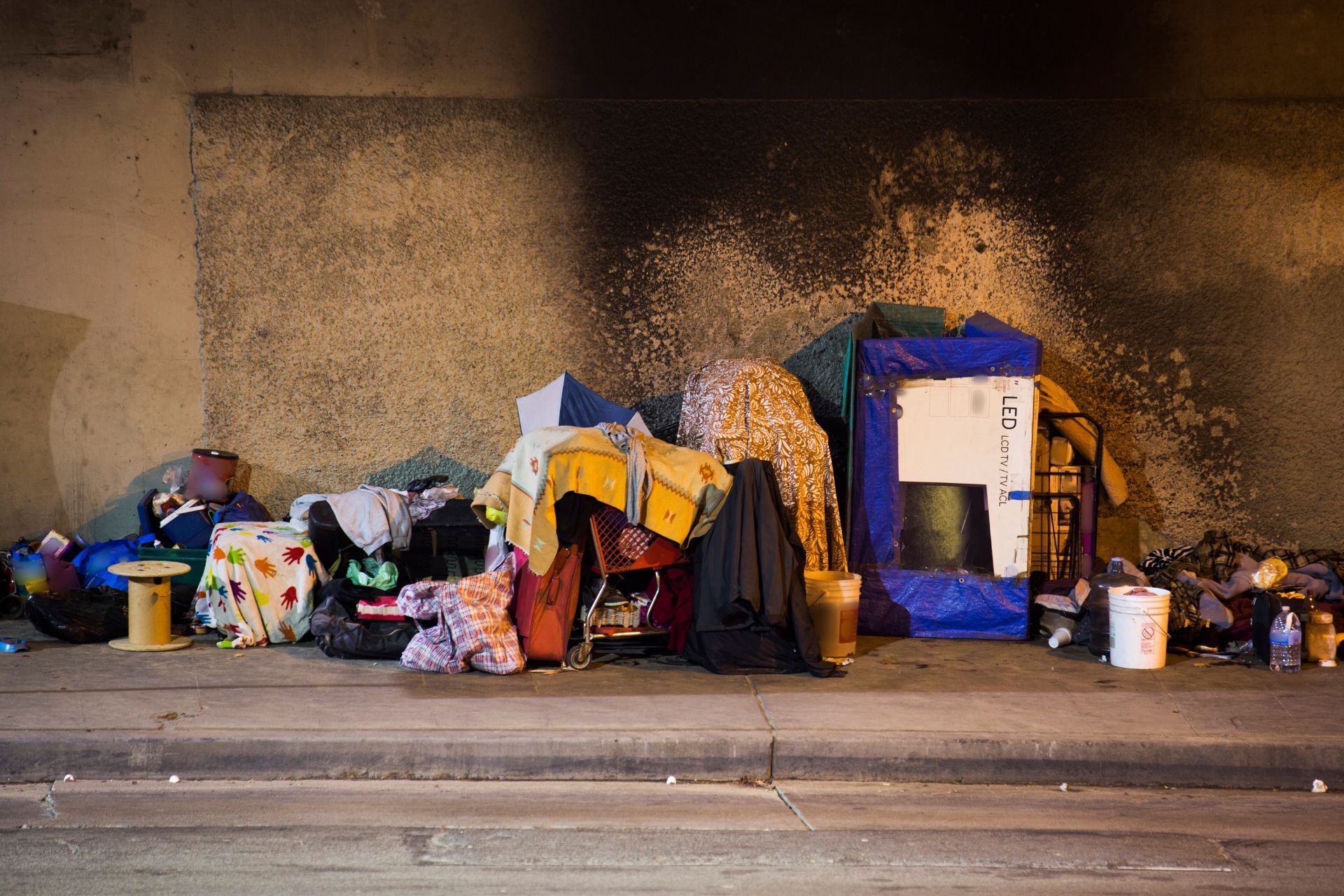 Pile of belongings on a sidewalk against a concrete wall, suggesting homelessness.