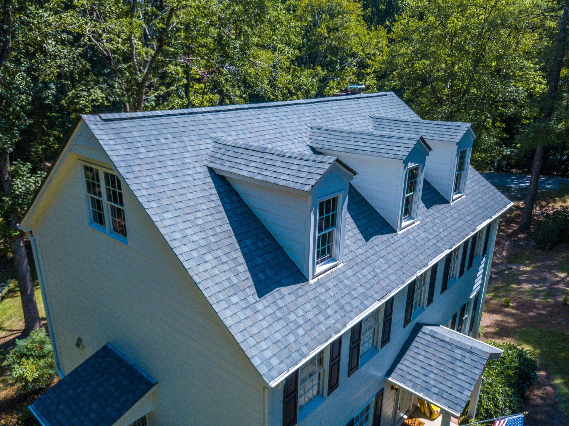 A two-story house with a gray shingled roof, three dormers, and black shutters, set amongst green trees.