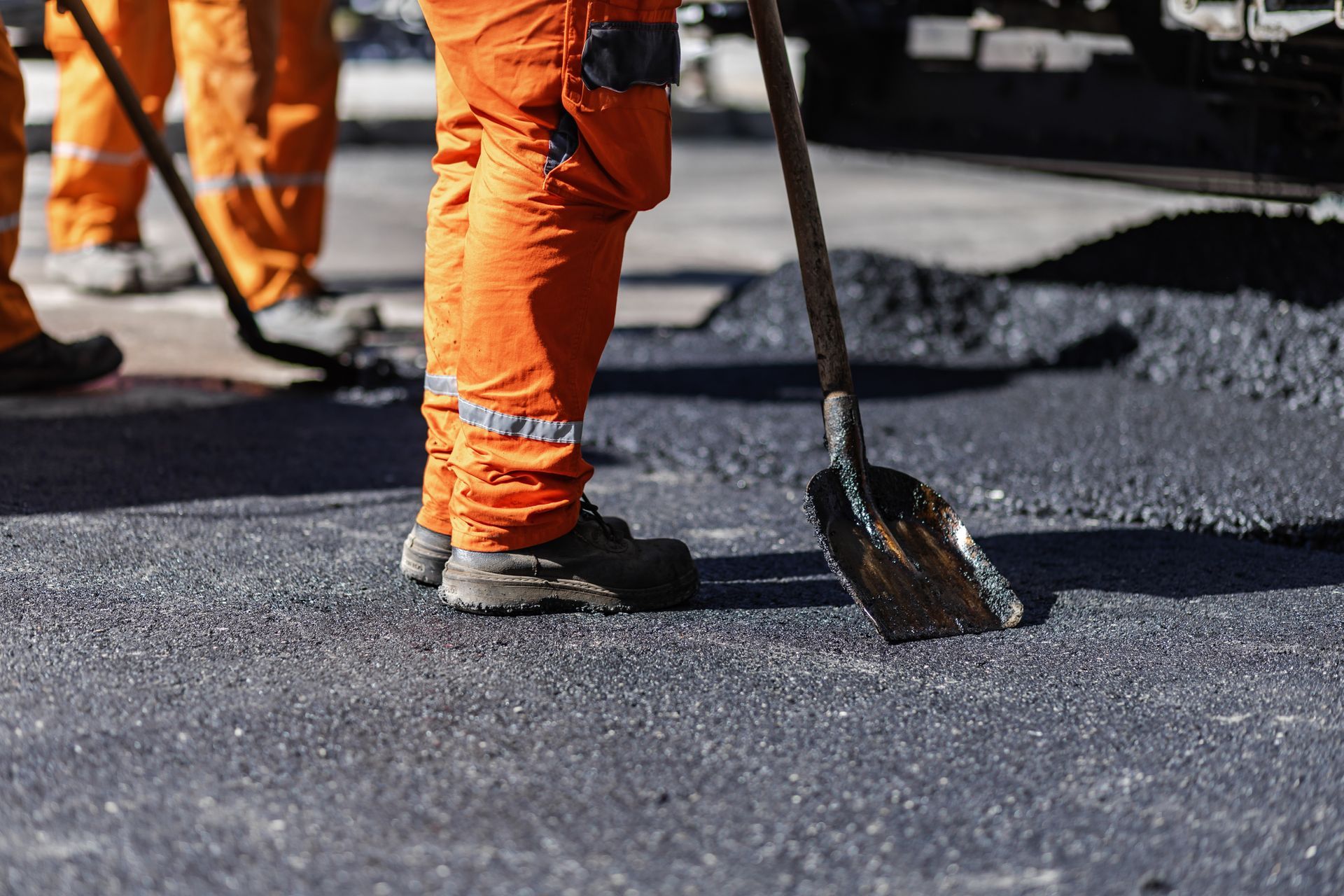 Construction workers in orange safety pants working on freshly paved black asphalt with a shovel.