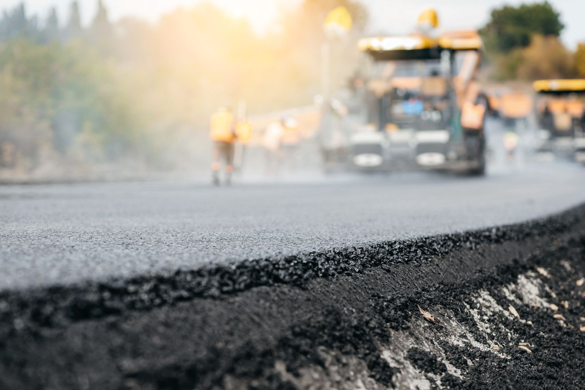 A construction vehicle paves a dark asphalt road with workers in the background at sunset.