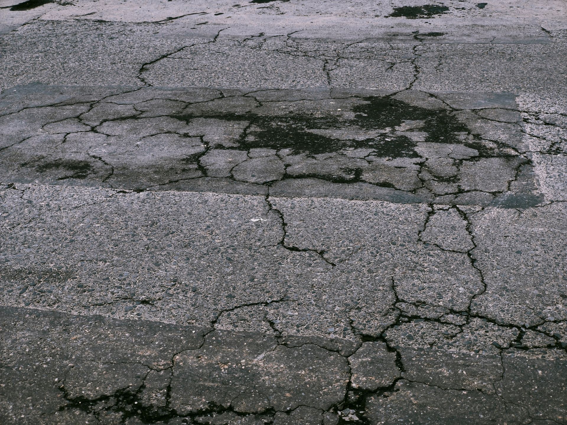 A close-up view of weathered, cracked gray asphalt pavement with dark patches and intersecting fissures.