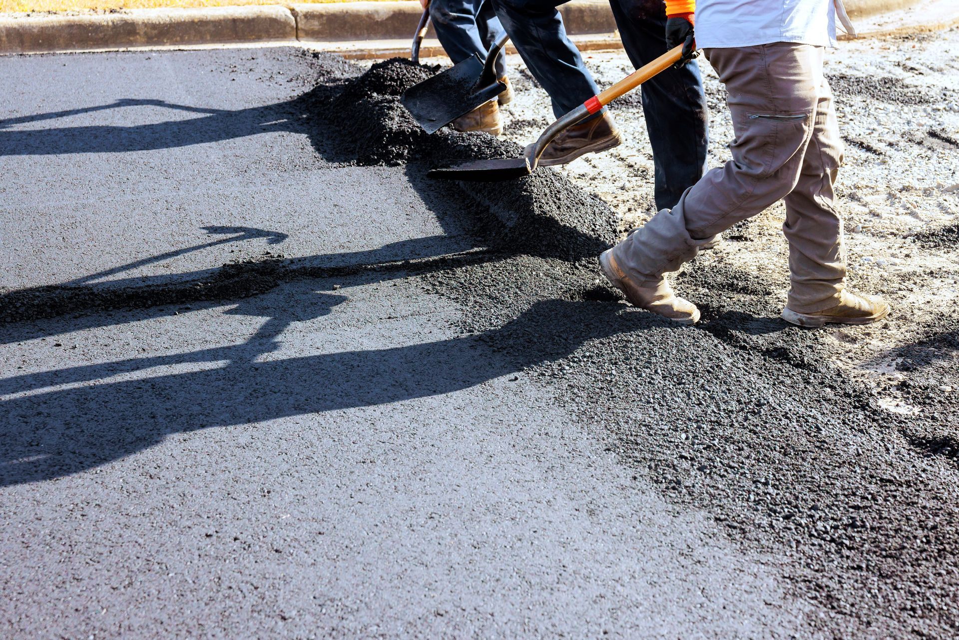 Workers use shovels to smooth fresh black asphalt onto a road surface.
