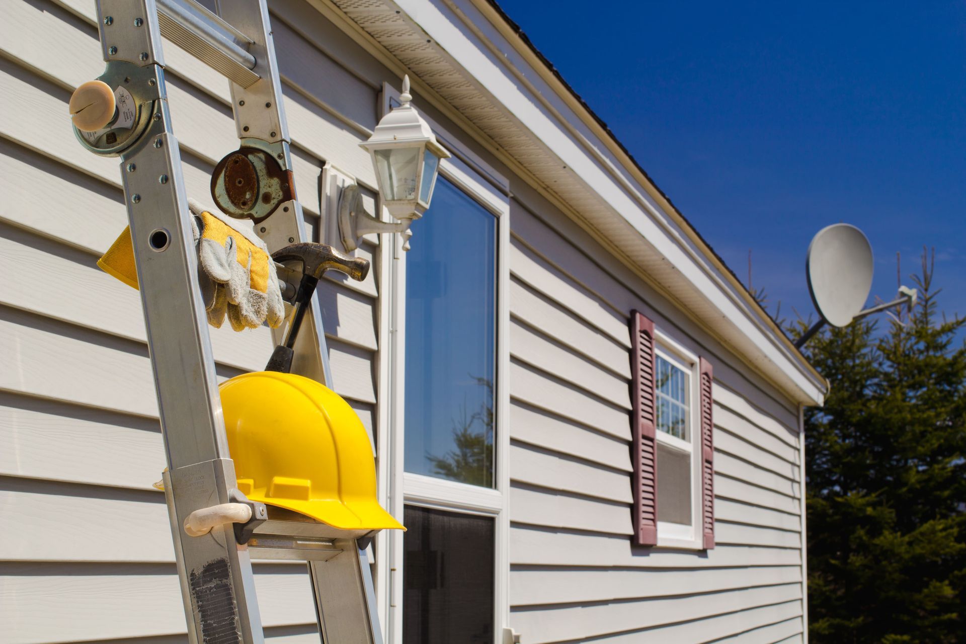 Yellow hard hat, gloves, and tools hanging on a ladder beside a house with white siding and a satellite dish.