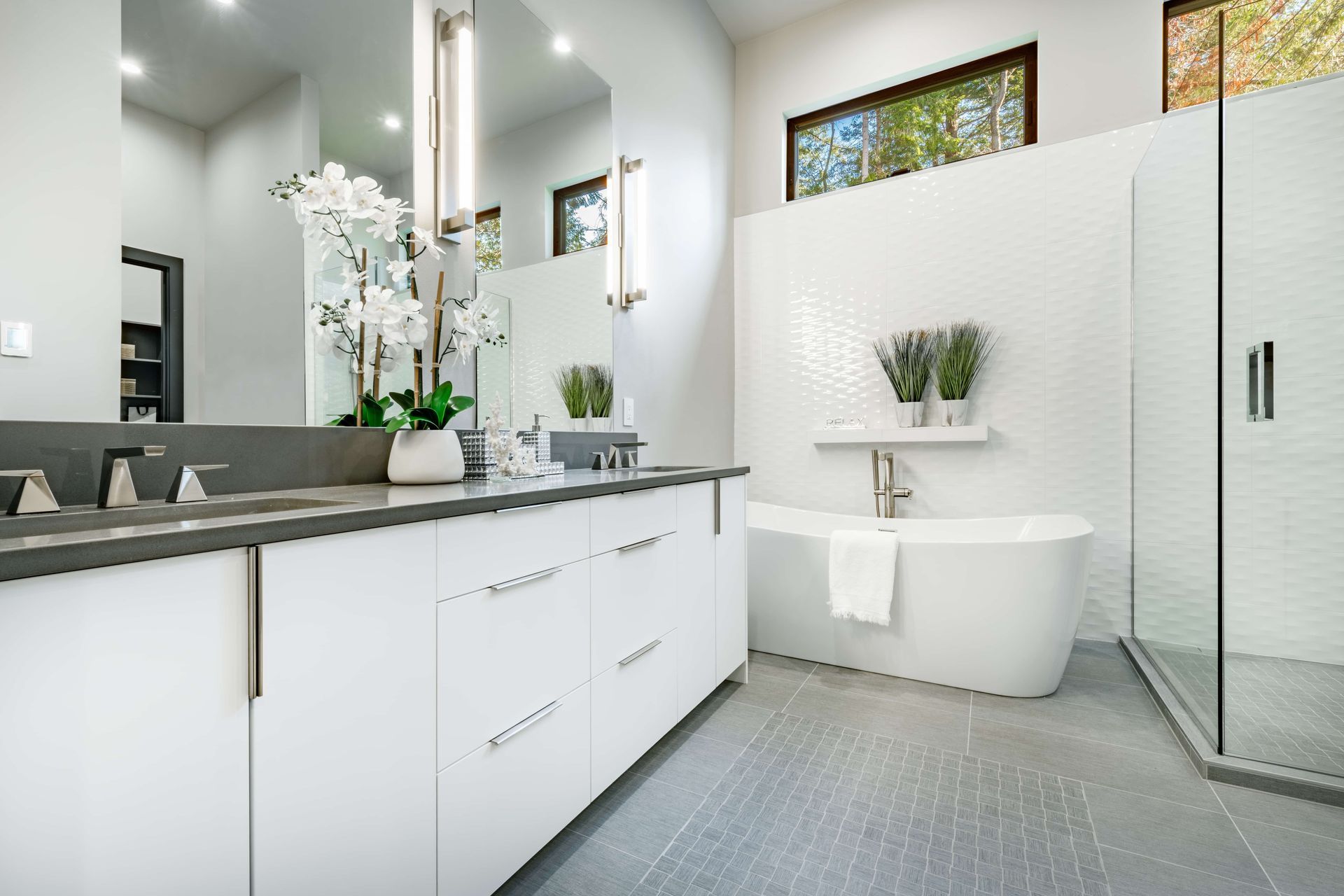 Modern white bathroom with gray countertop, freestanding tub, glass shower, and decorative plants.