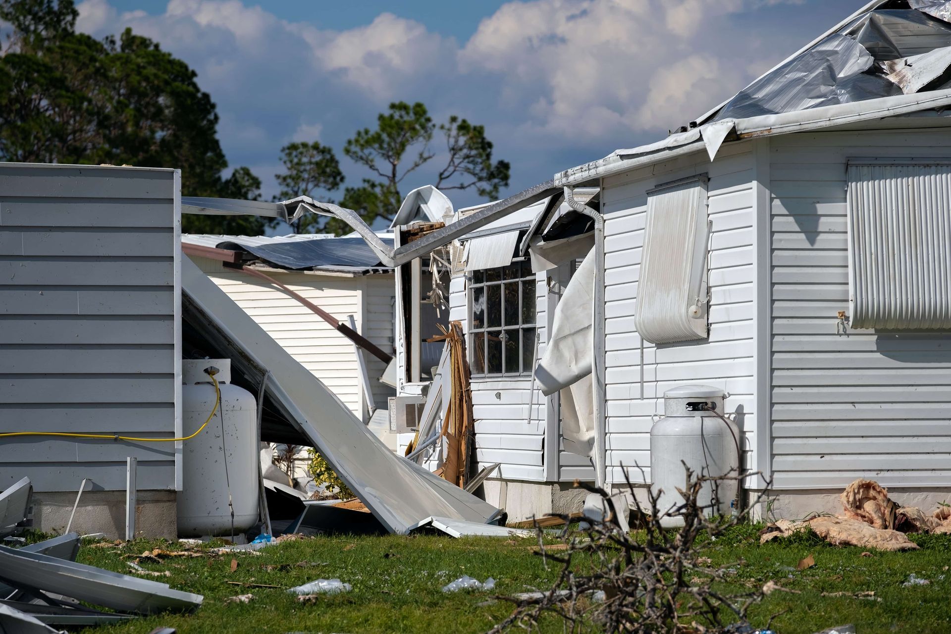 House heavily damaged by a storm; white siding torn away, debris scattered on green grass under a blue sky.