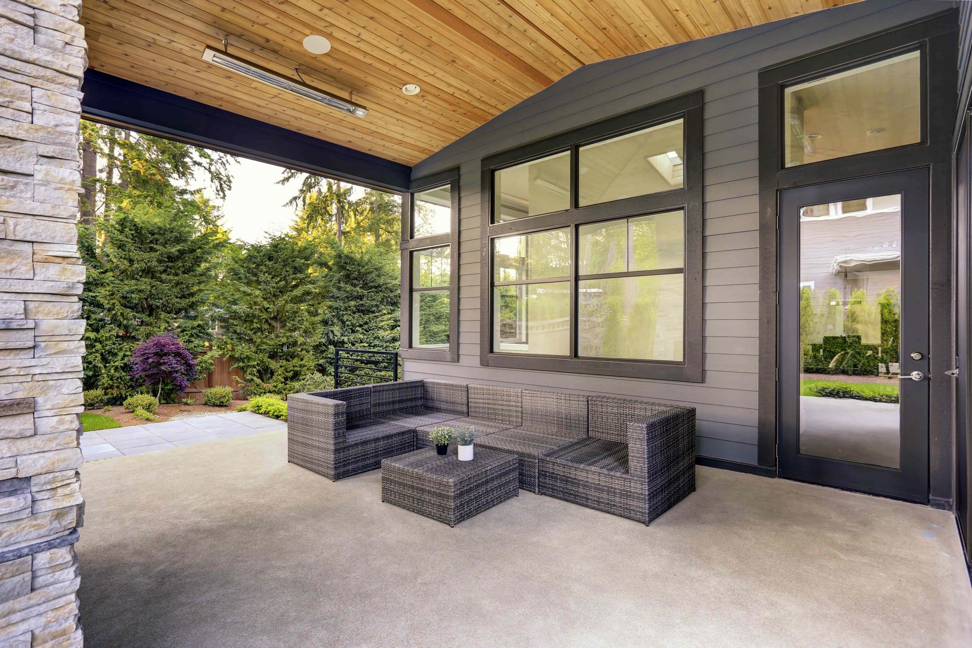 Outdoor patio with gray wicker sofa and coffee table, wooden ceiling, and lush greenery.