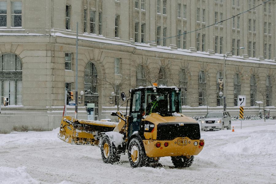 Yellow snowplow clearing snow on a city street in front of a large building, snowy weather.