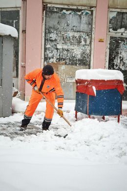 Man in orange jumpsuit shoveling snow in front of a building with faded blue doors.