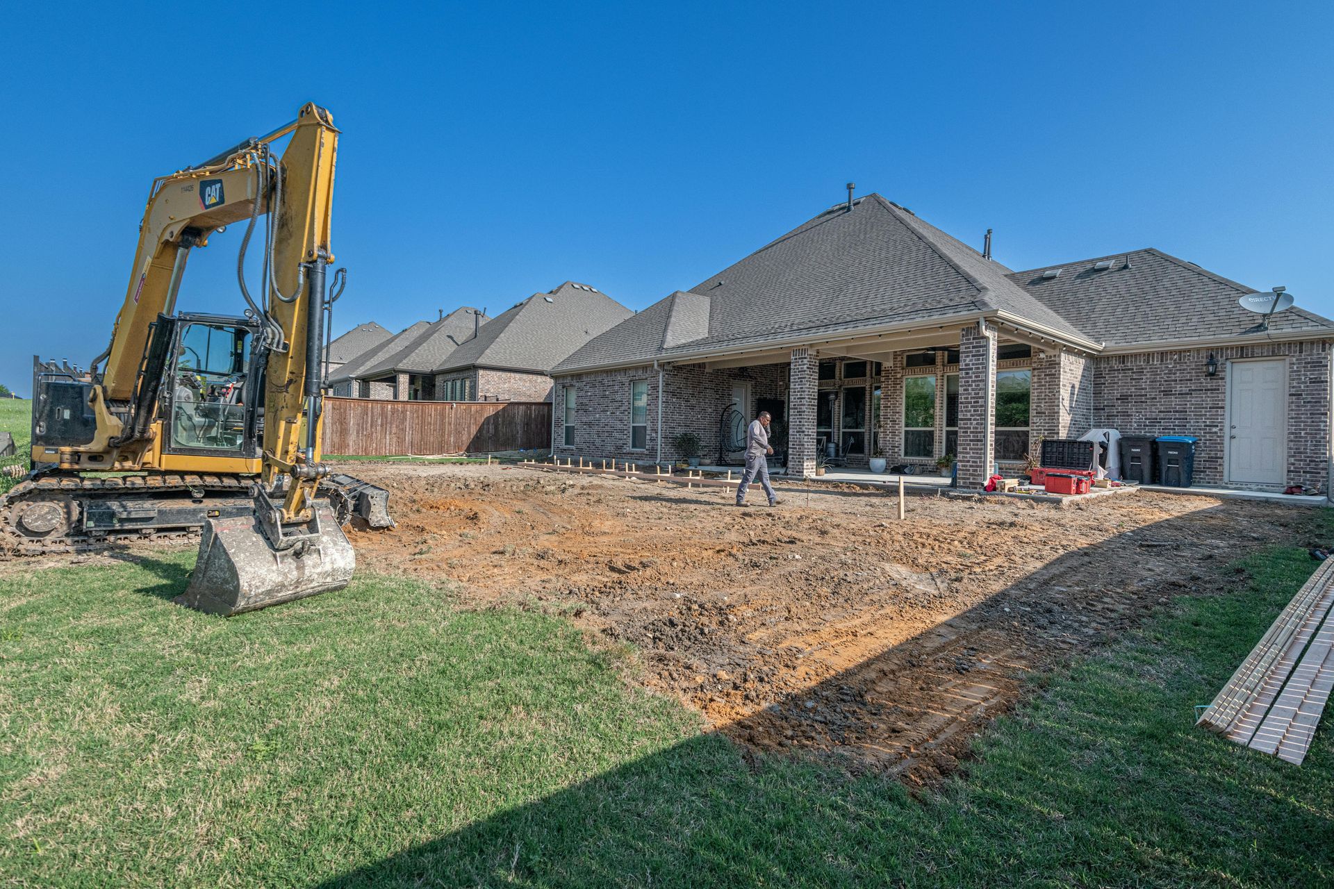Yellow excavator on a sunny day, working on a backyard patio near a brick house.