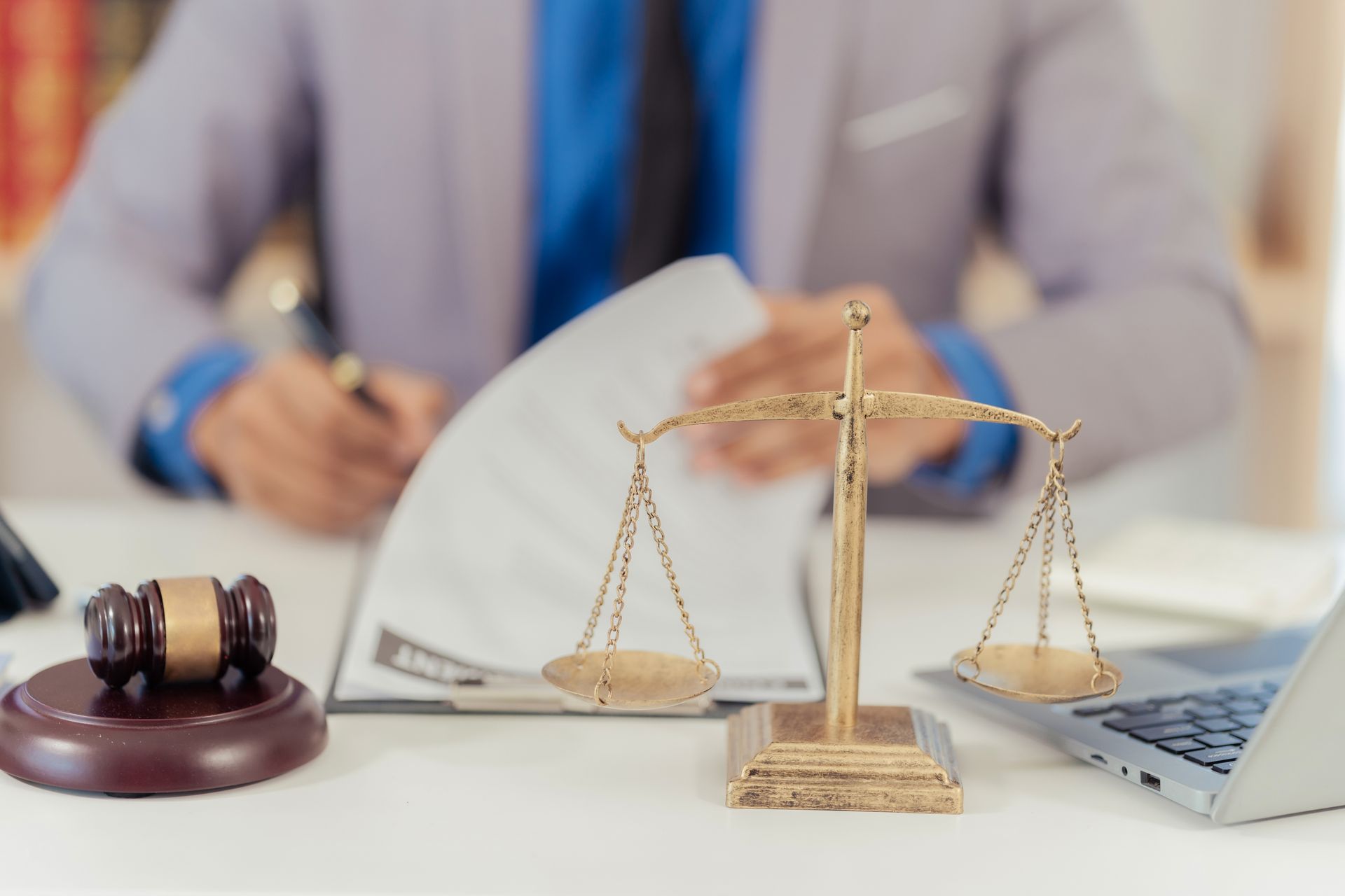 A lawyer sits signing a document at a desk with a laptop, a gavel, and the Scales of Justice
