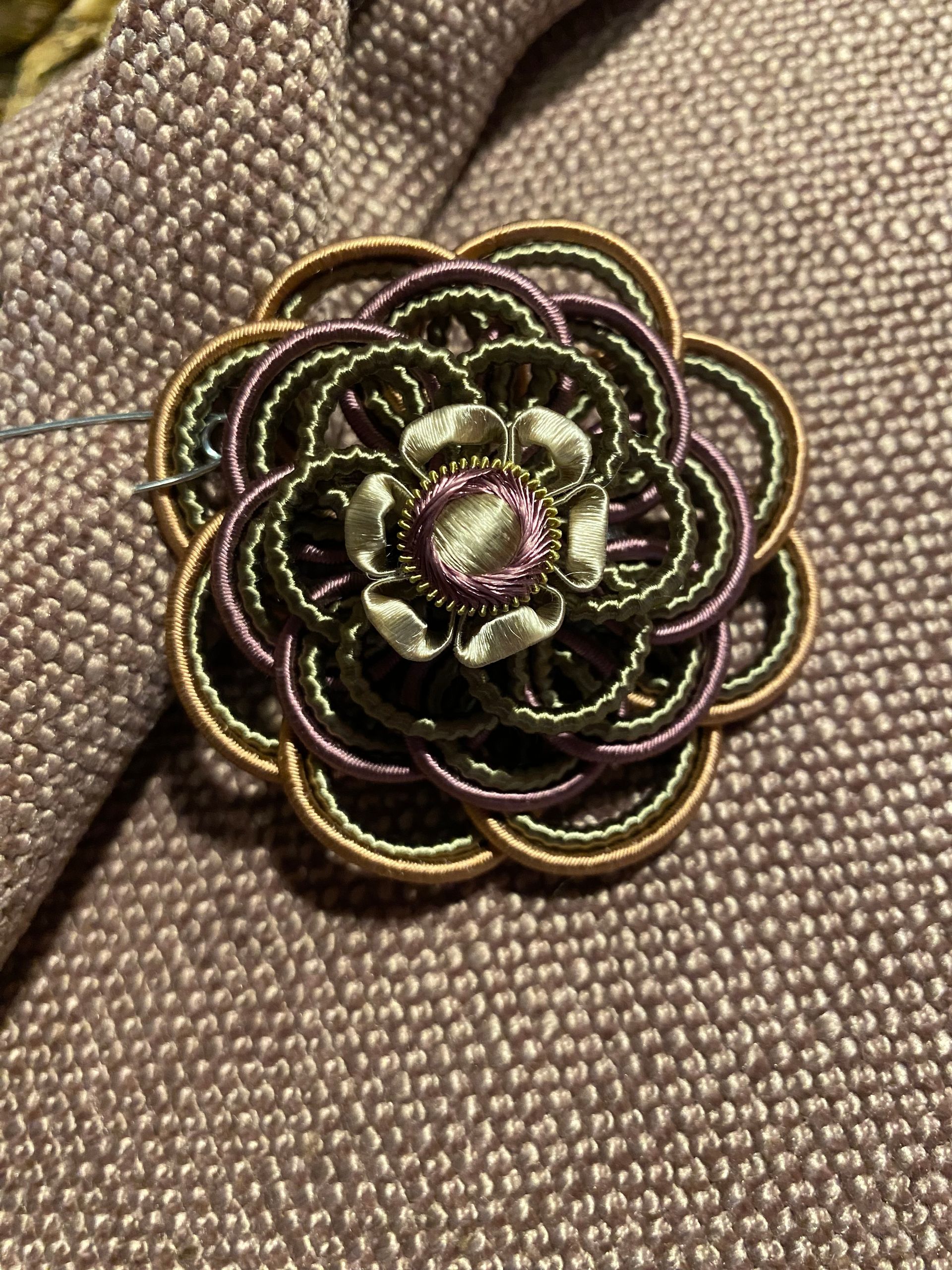 Brooch shaped like a flower, with concentric circles of gold, purple, and green thread, pinned to a mauve textured fabric.