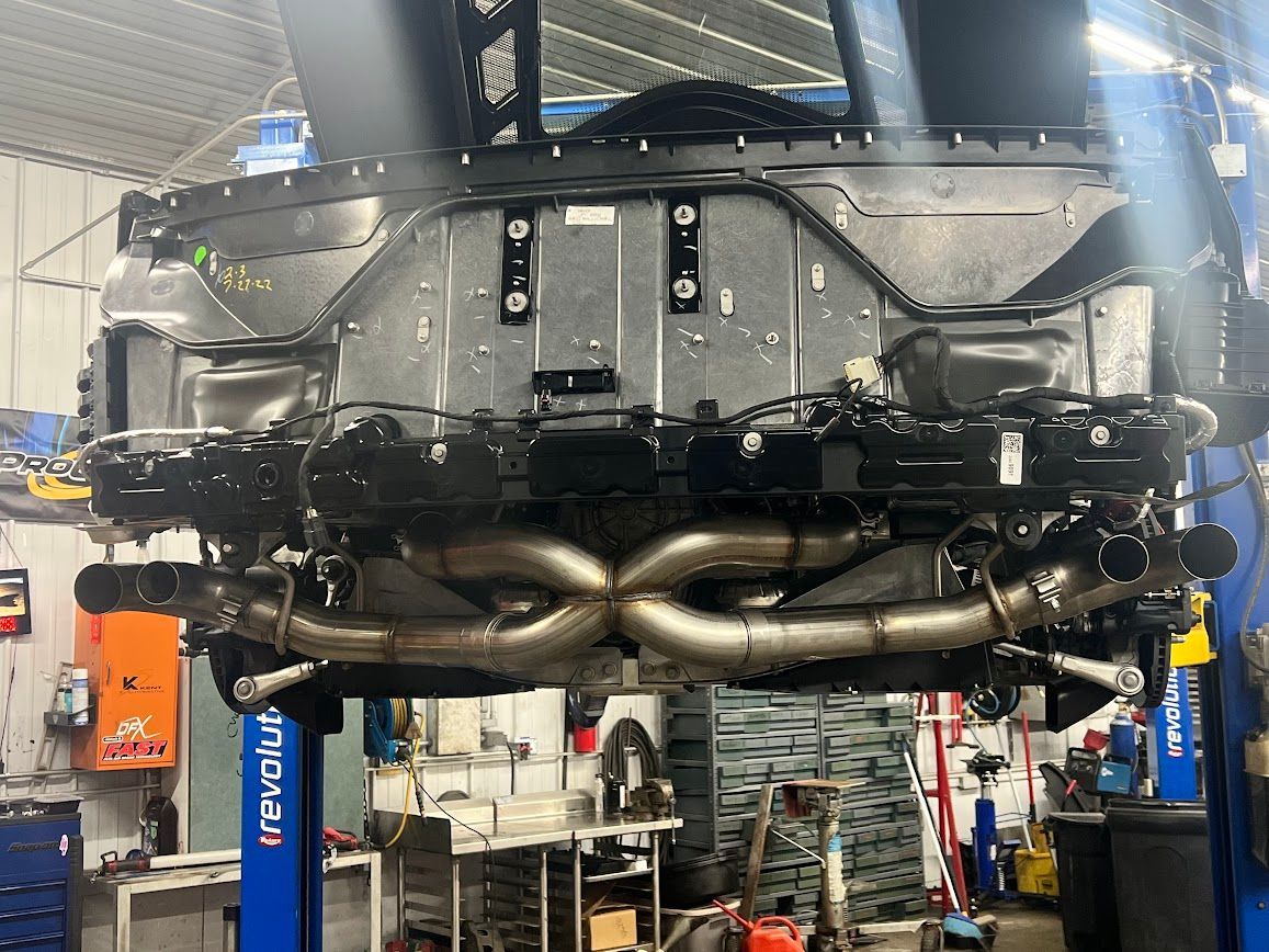 Underside of a car on a lift; silver exhaust pipes, black chassis, repair shop setting.