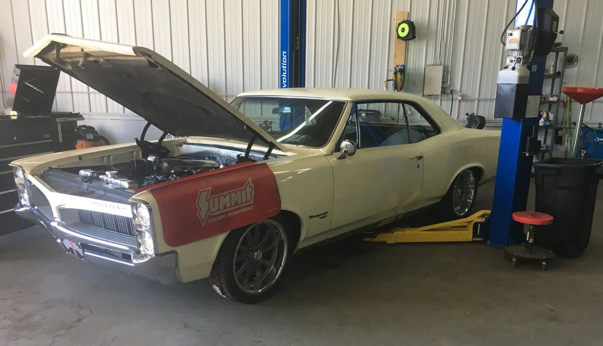 Cream-colored classic car with hood open in auto shop, on a lift, with a red fender cover.