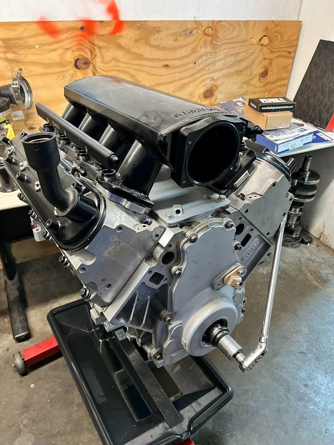 An engine block, with intake manifold, in a garage setting. It's gray with black intake and a pulley.
