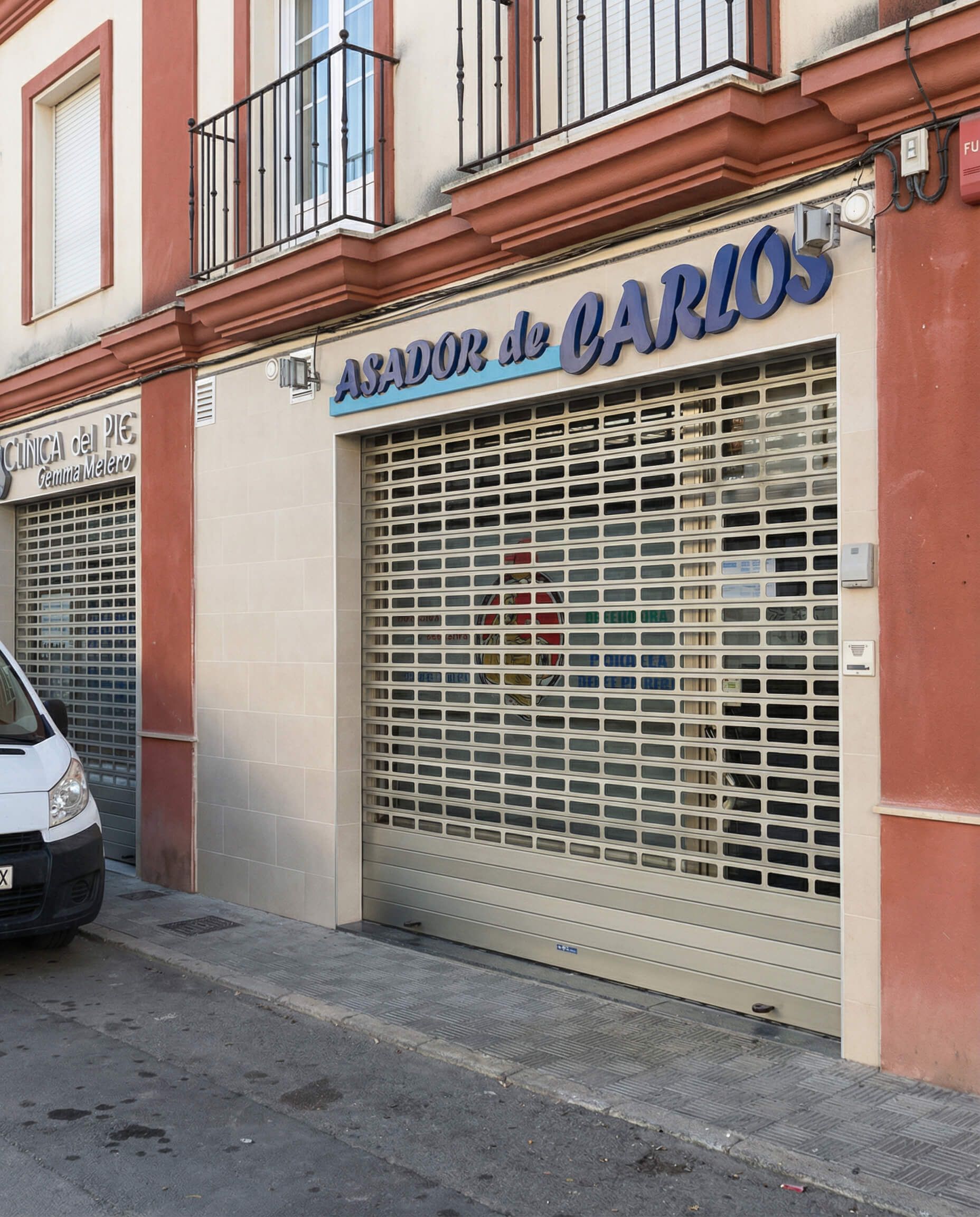 Fachada de edificio con cartel de "Asador de Carlos" encima de persiana metálica de seguridad cerrada.