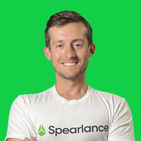 A man wearing a white spearlance t-shirt is smiling with his arms crossed.