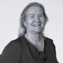 Smilende person med langt hår iført mørk skjorte og briller hengende fra kragen