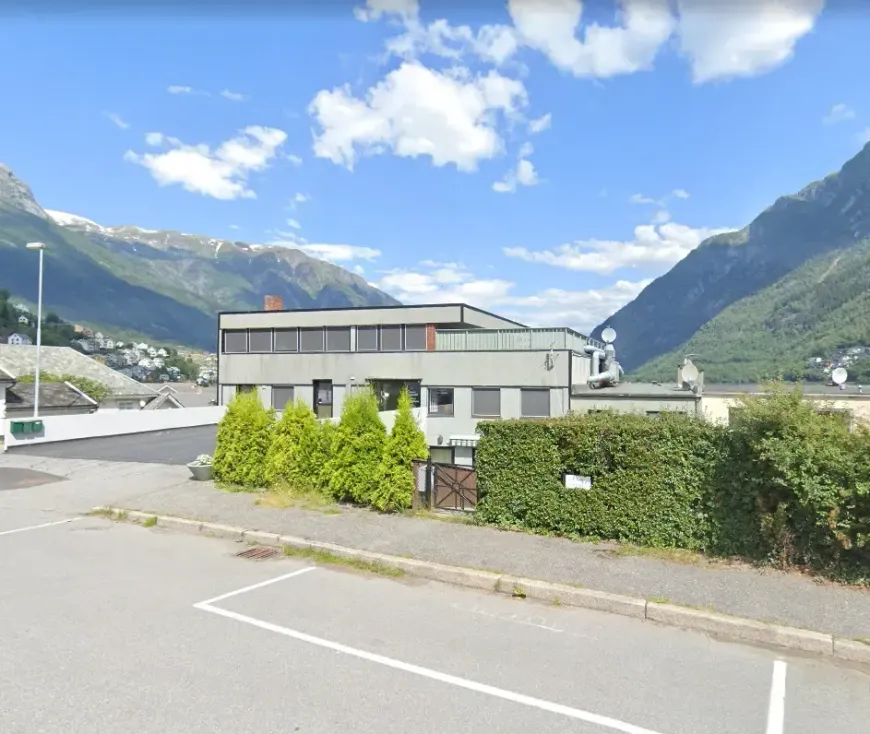 Fjelllandsbygate med et grått hus, grønne hekker og snødekte topper under en blå himmel