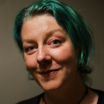 Nærbilde av en person med blågrønt hår, nesering og et svakt smil mot en mørk bakgrunn