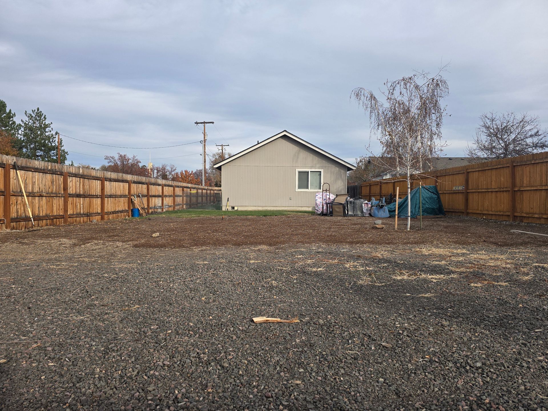 A backyard with a gravel foreground, a tan shed, and a wooden fence under an overcast sky.