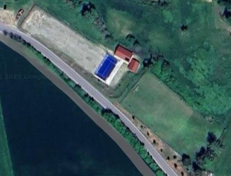 Vista aerea: complesso sportivo con un campo blu, un prato verde ed edifici rossi accanto a una strada e a uno specchio d'acqua.