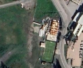 Vista aerea di un campo da tennis accanto a un cantiere e a una strada; circondato da erba e altri edifici.