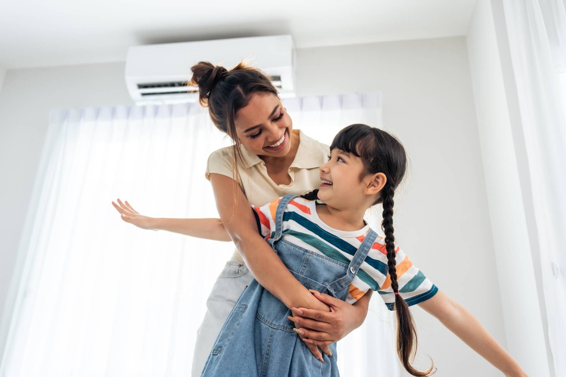 Woman hugging smiling child in a bright room with white curtains and an air conditioner.