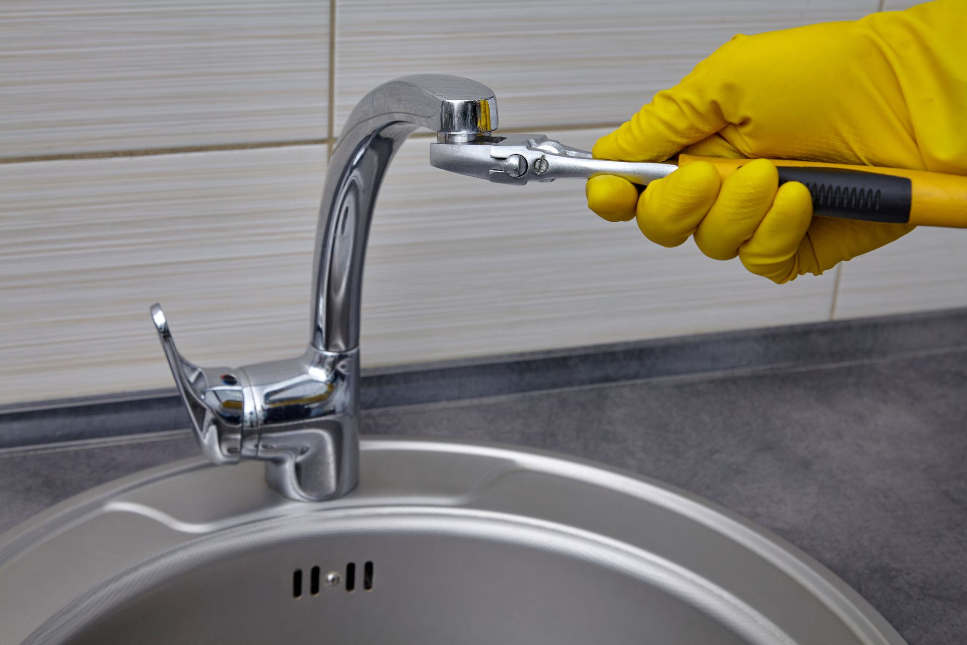 A person wearing yellow gloves is fixing a sink faucet with a wrench.