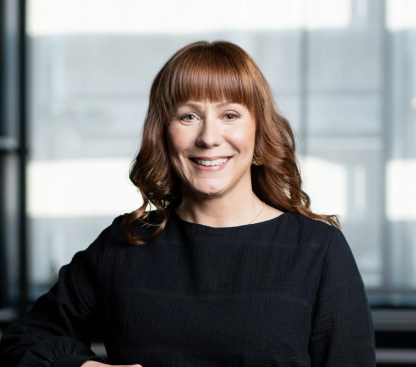 Woman with reddish-brown hair and bangs smiles in a modern office. She wears a black top.