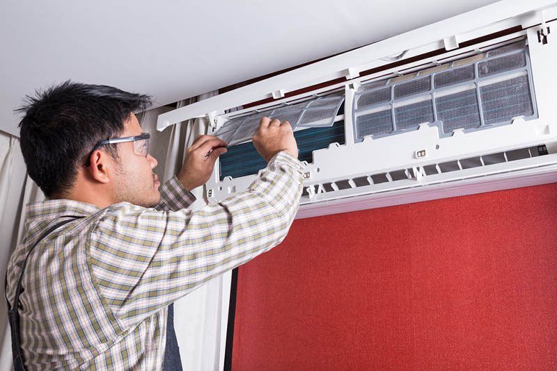 Person in glasses cleaning an air conditioner filter in a room with a red wall.