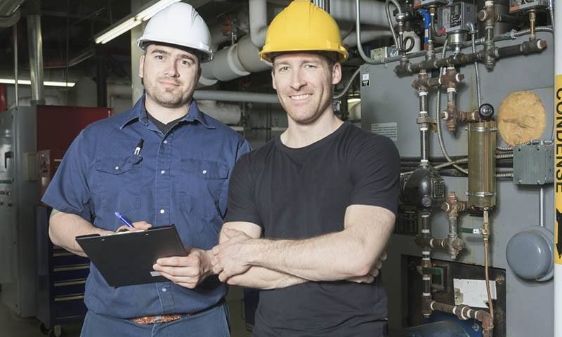 Two workers in hard hats pose in a machinery room. One holds a clipboard; the other has crossed arms.