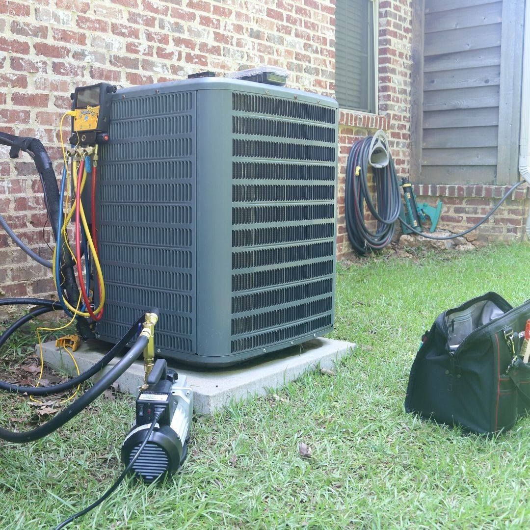HVAC unit being serviced outdoors, with tools, hoses, and vacuum pump on grass next to a brick building.