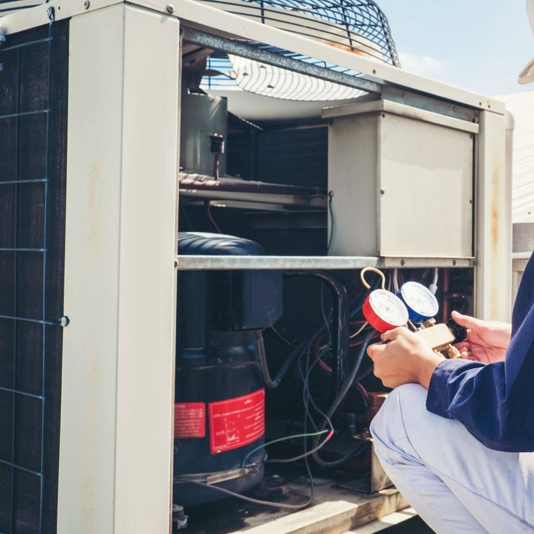 HVAC technician kneels, checking gauges on an open rooftop air conditioning unit.
