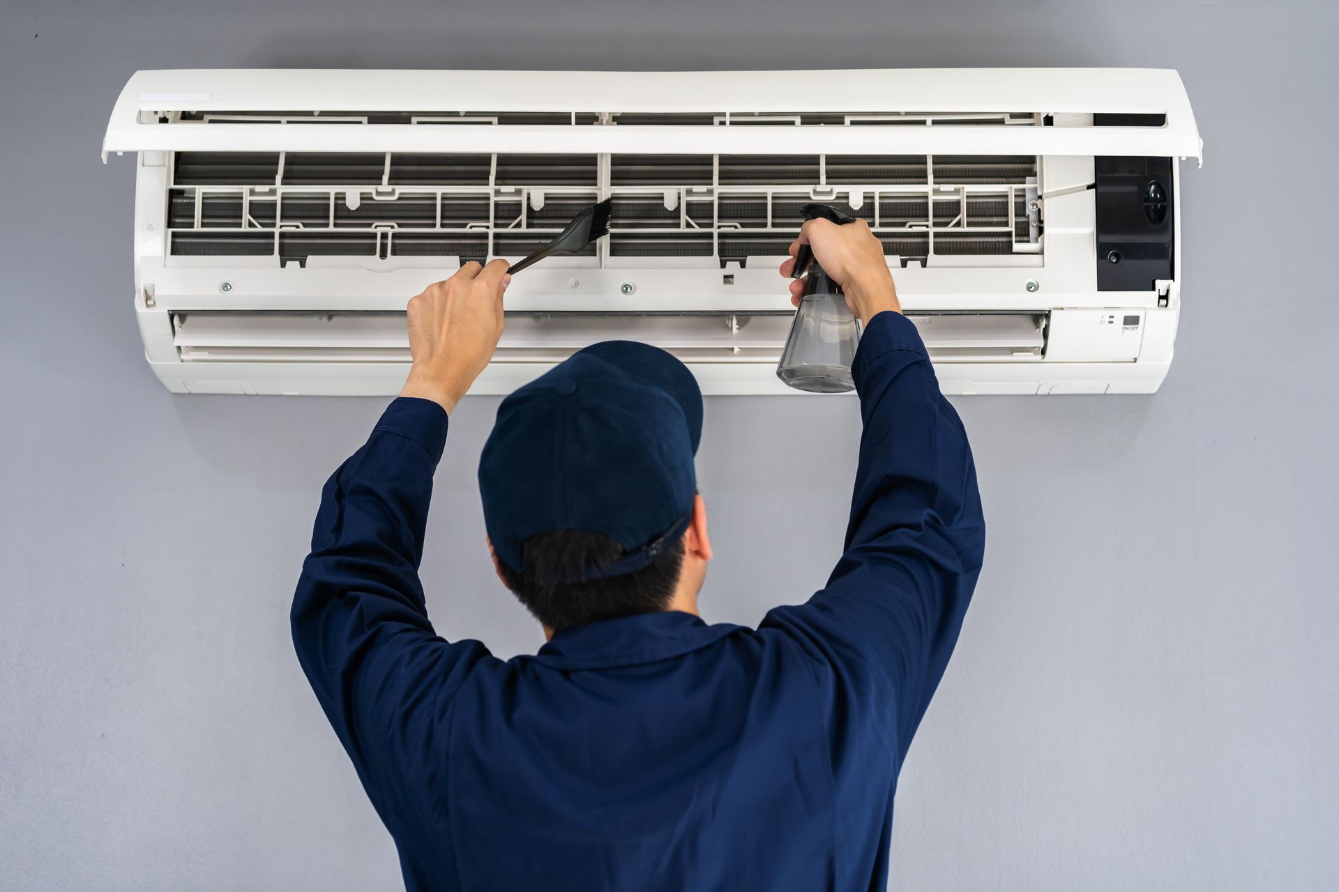 Person in blue uniform cleaning a white air conditioning unit with a brush and spray bottle.