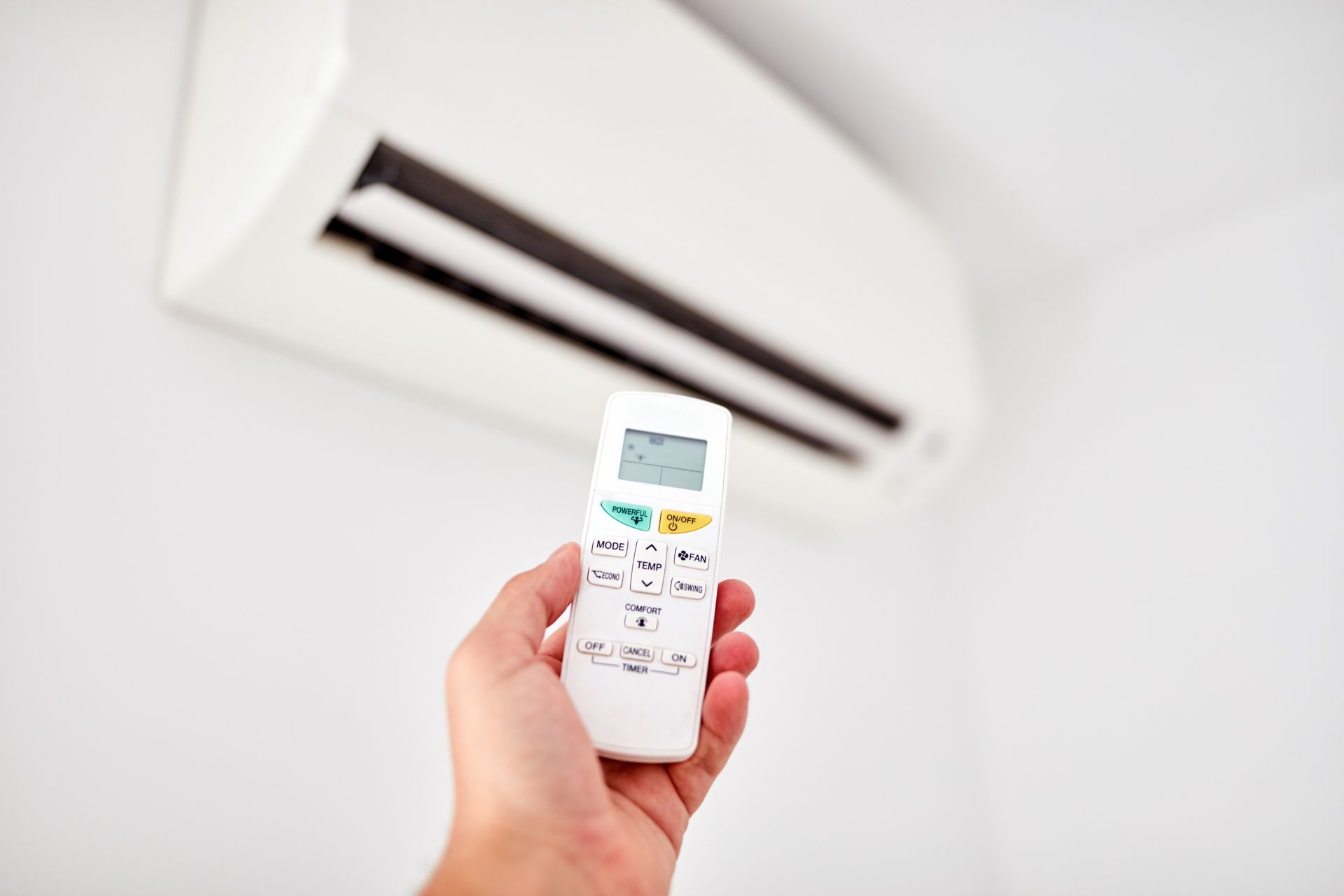 Hand holding air conditioner remote, aimed at white wall-mounted unit.