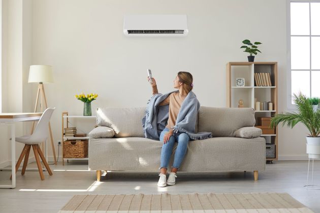 Woman on sofa adjusts AC with remote in a bright living room.
