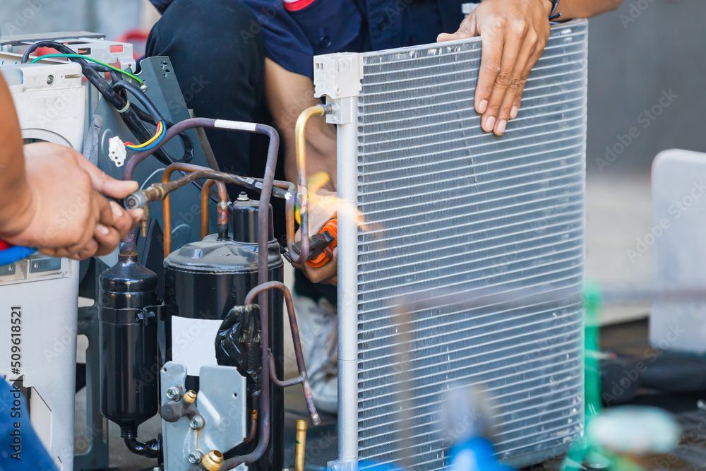 Two technicians repairing an air conditioning unit, one holding a radiator.