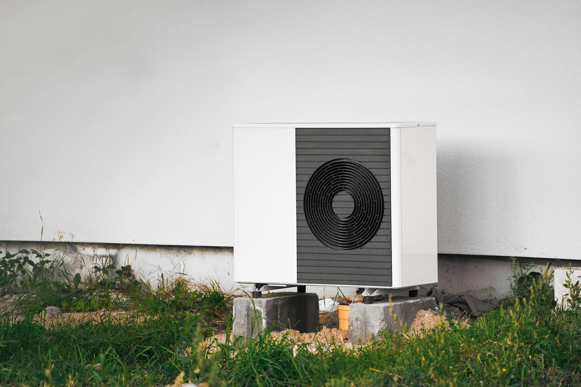 White rectangular heat pump unit mounted on a concrete block, against a white wall and overgrown weeds.