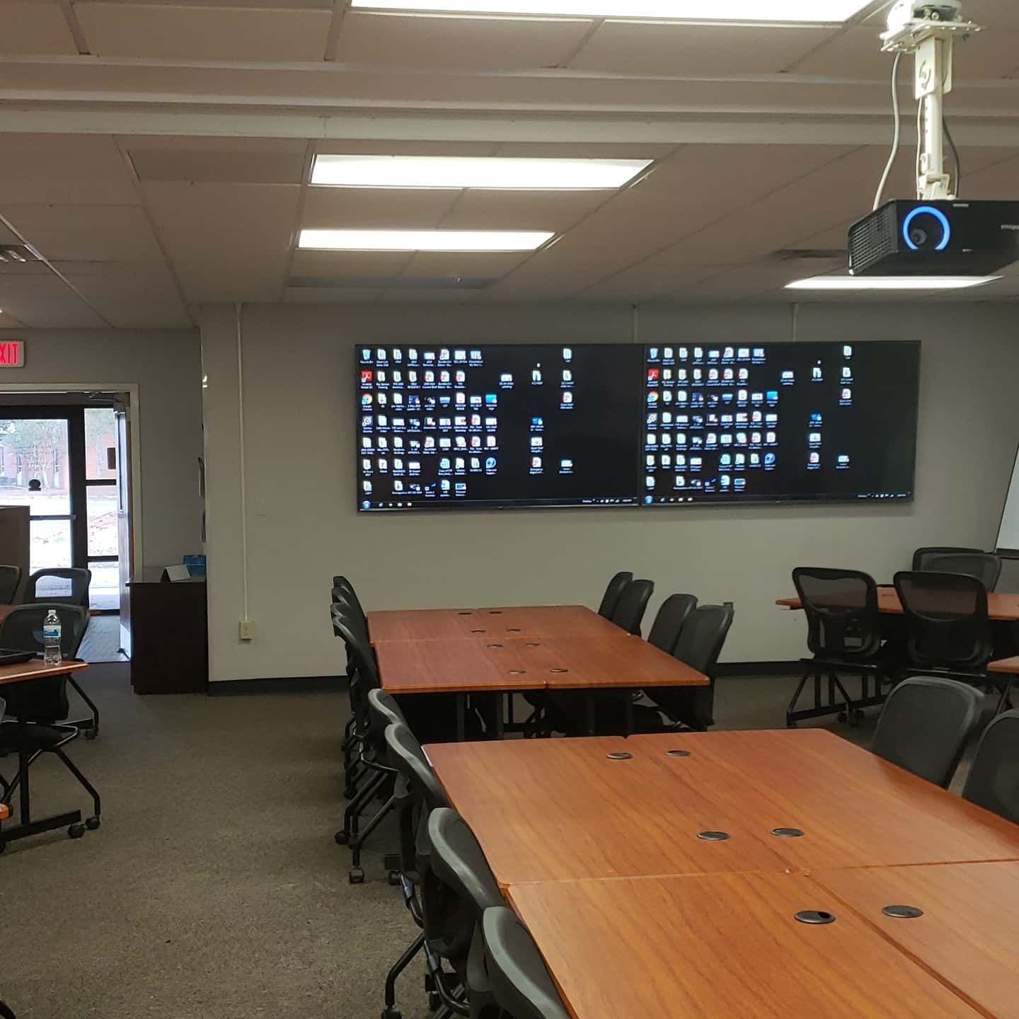 Empty classroom or conference room with long tables, black chairs, wall-mounted screens, and a projector
