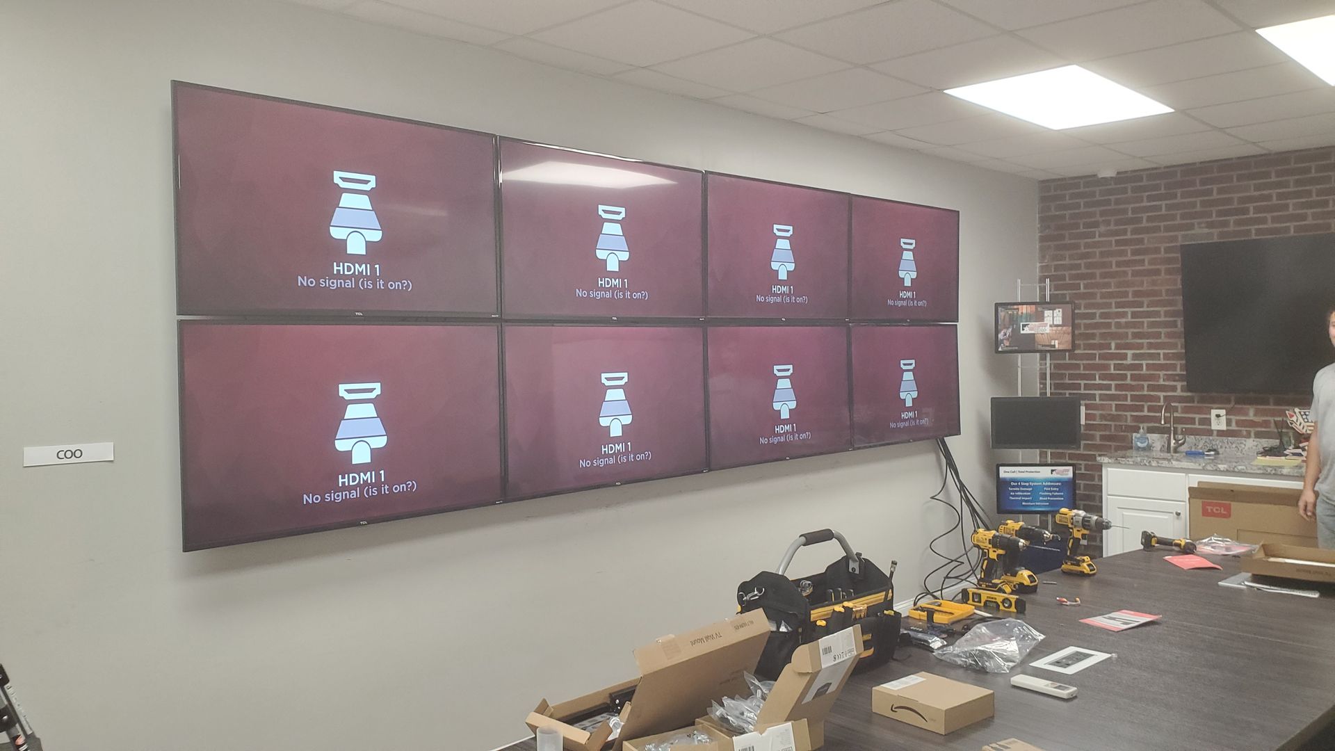 Large wall of eight monitors in a workshop or office, with equipment and boxes on a table below.