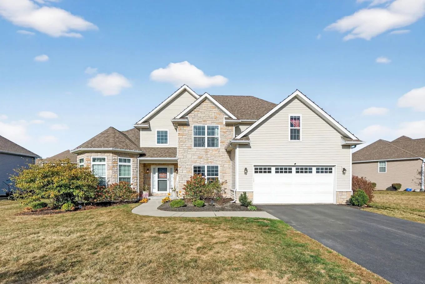Two-story beige house with stone accents, attached garage, and a lawn under a blue sky.