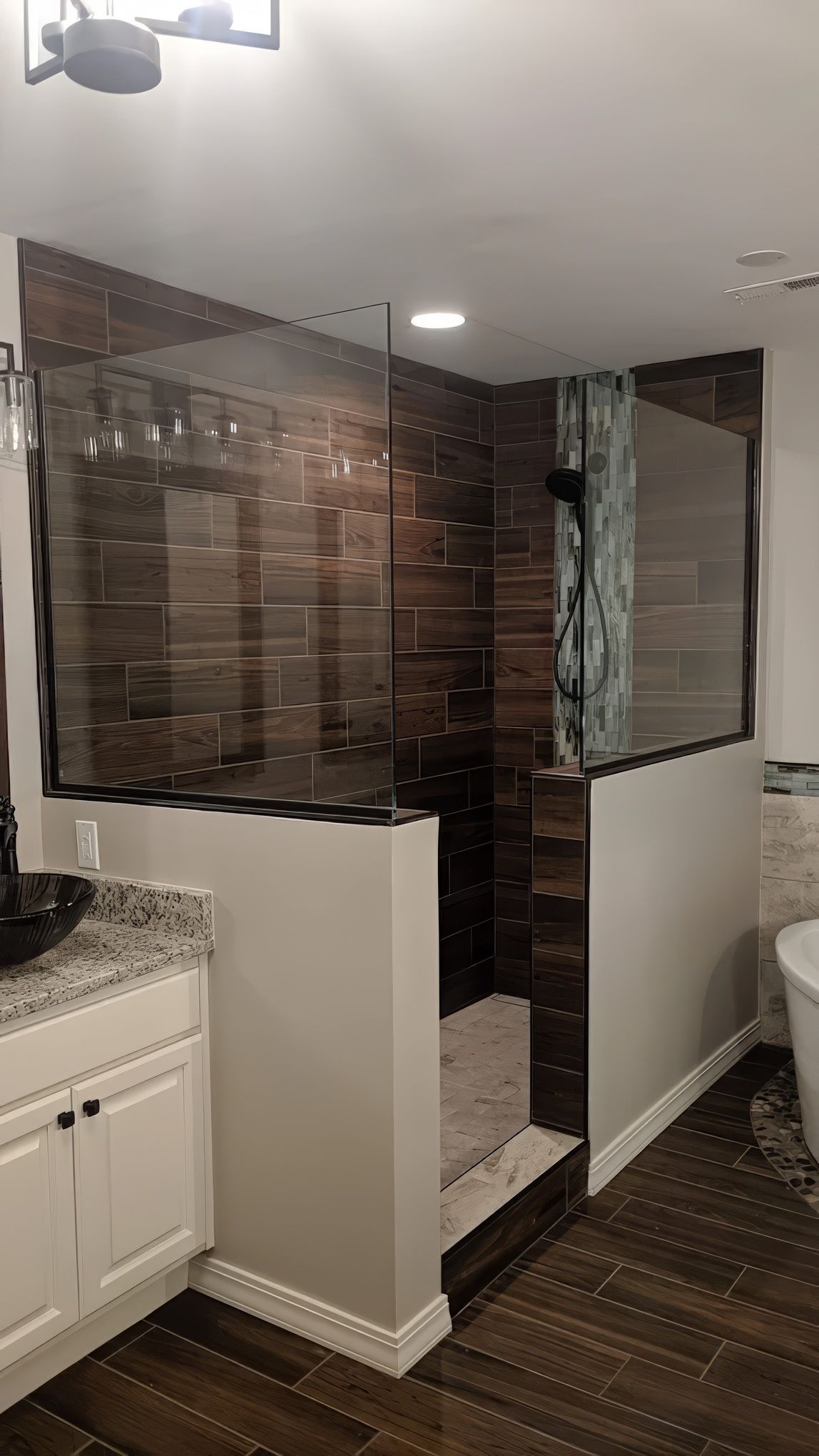 A walk-in shower with brown wood-look tile, glass walls, and a white vanity in a modern bathroom.