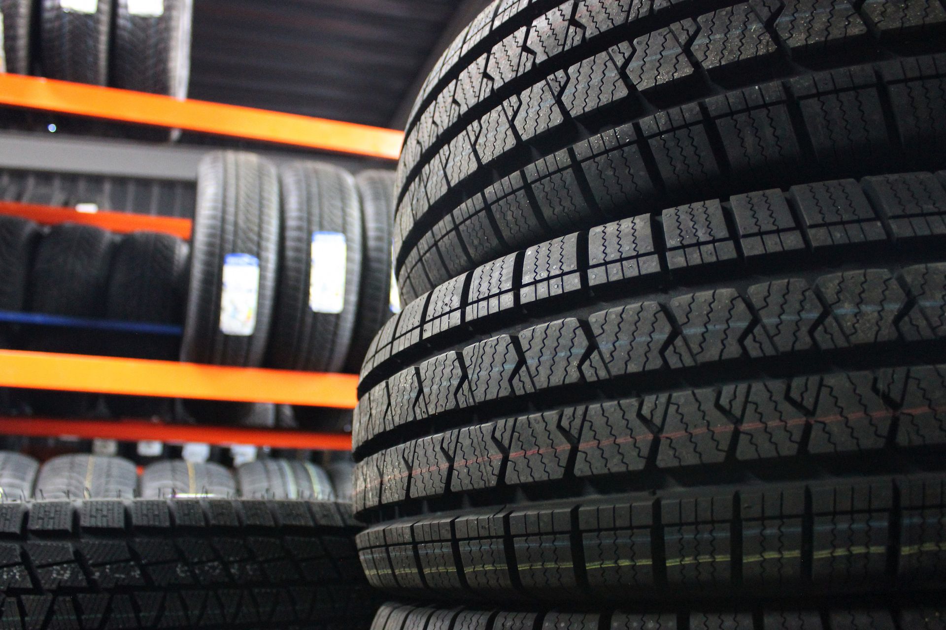 A stack of new, black winter tires in the foreground with rows of identical tires on orange warehouse shelving behind.