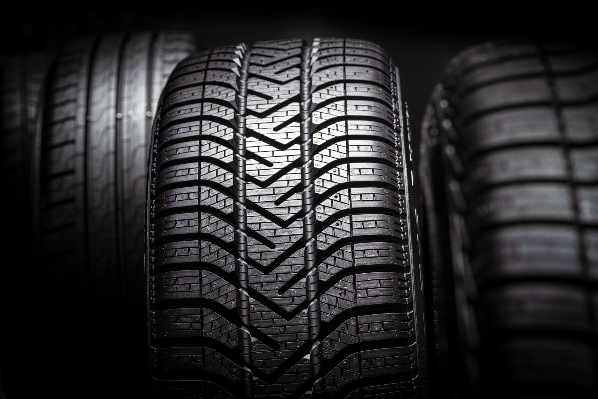 Close-up of a dark, textured car tire tread in the center, flanked by two other tires in a dark, moody background.